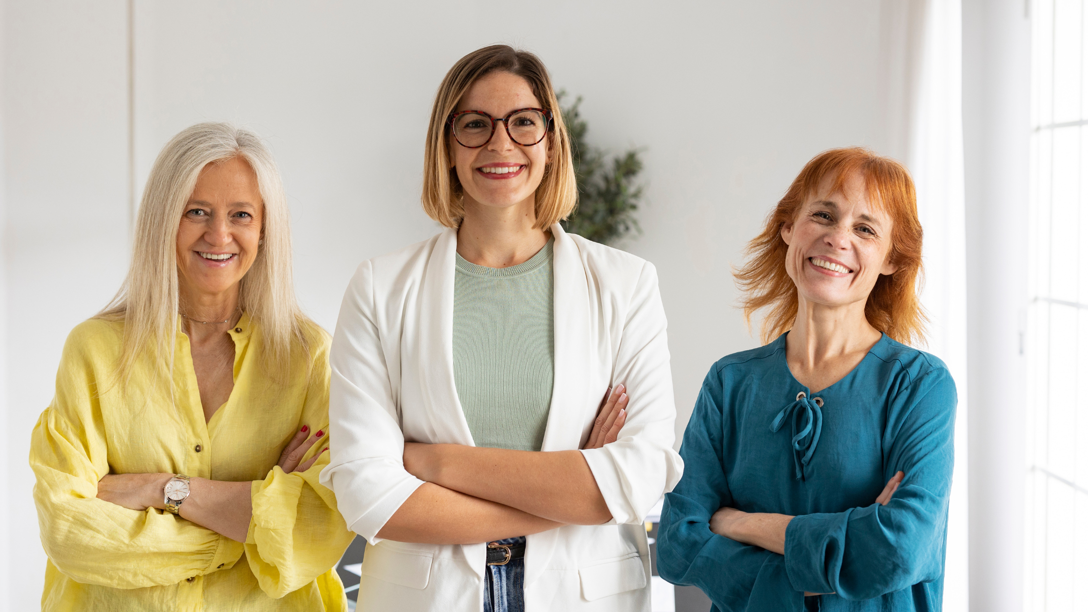 Untitled design (12) Three women standing. The left has yellow shirt on middle has white shirt on and right has a blue shirt on